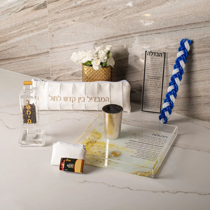 Square brass besamim holder with spice container and blue braided cover, displayed with Shabbat essentials on white surface.