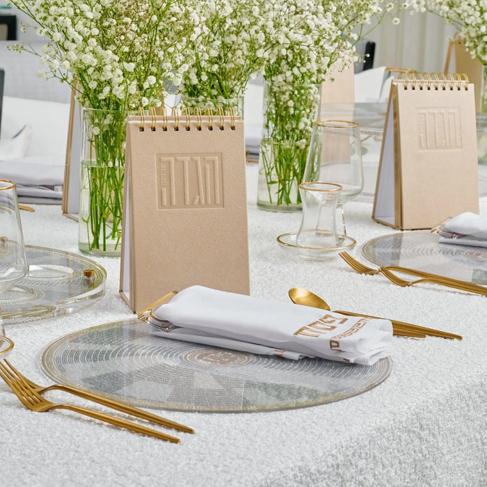 Standing leather Haggadah in tan with gold accents on a styled Pesach table setting with white linens and greenery.