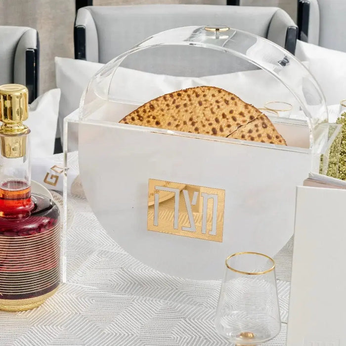 White lucite standing matzah box with suspended compartment, displayed on tabletop with Pesach items and cosmetics nearby.