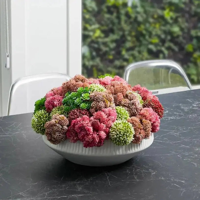 Pink and white faux flower succulent arrangement in a ceramic white pot on a dark surface with greenery backdrop.