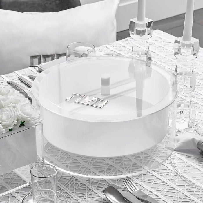 Suspended matzah box in clear lucite displayed on a bathroom counter with glass accessories and white towels nearby.