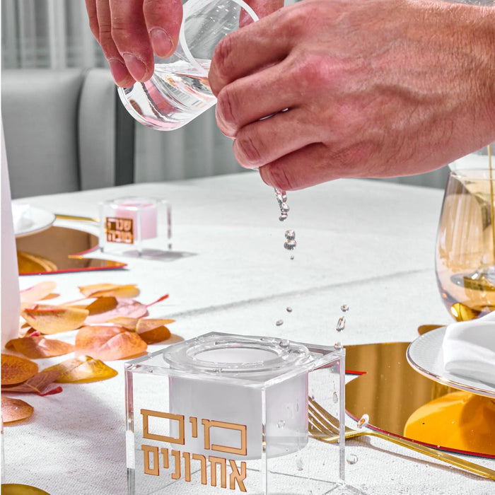 Clear glass suspended mayim achronim dispenser with golden liquid, displayed on white table with place settings and flowers.