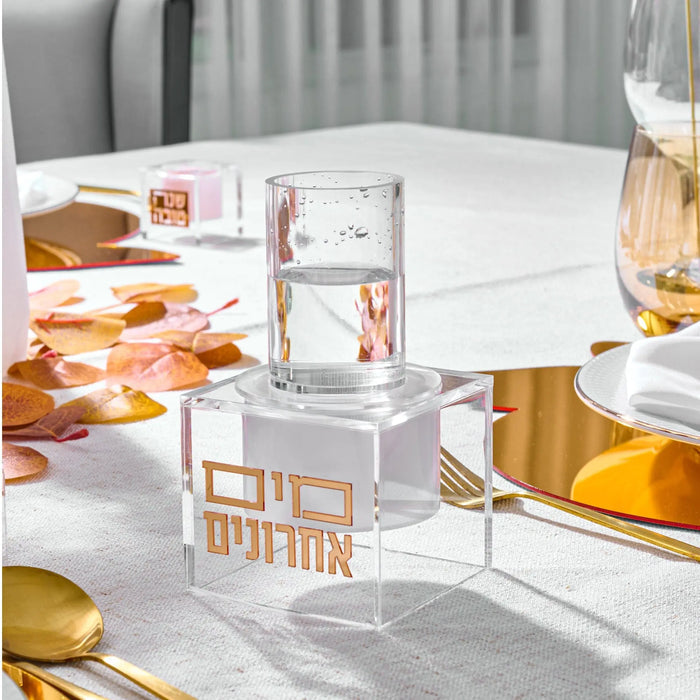 Clear acrylic suspended mayim achronim dispenser on white table with gold accents and place settings, perfect for elegant Shabbat and holiday entertaining.