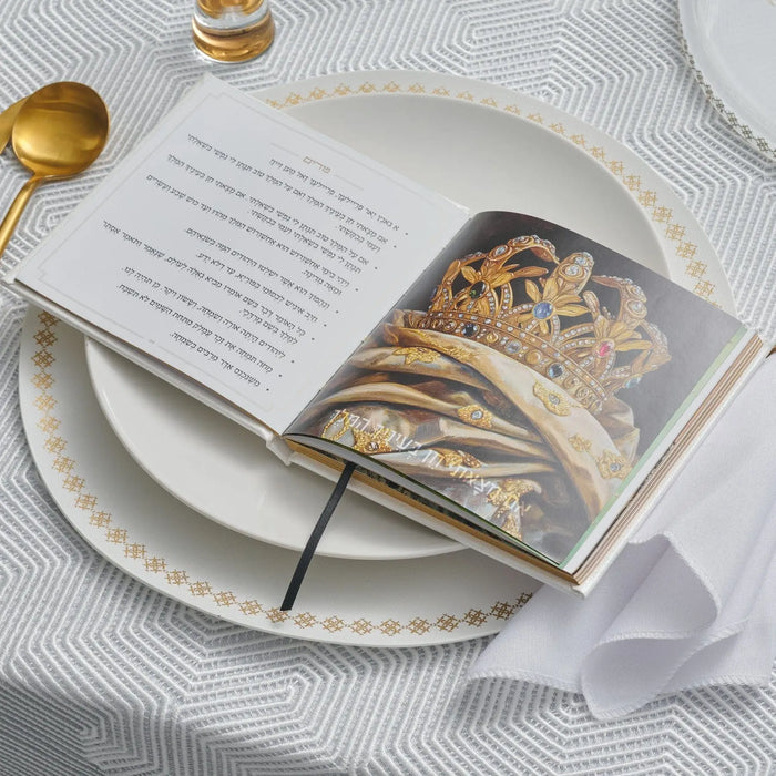 White plate setting with cream and gold Suspended Shabbos bencher, gold utensils, and prayer book for Shabbat meals and blessings.
