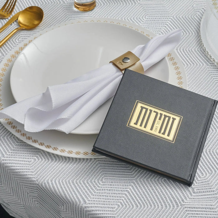 Black and gold Shabbos bencher set with white napkin and gold utensils on textured table setting for Shabbat meals.