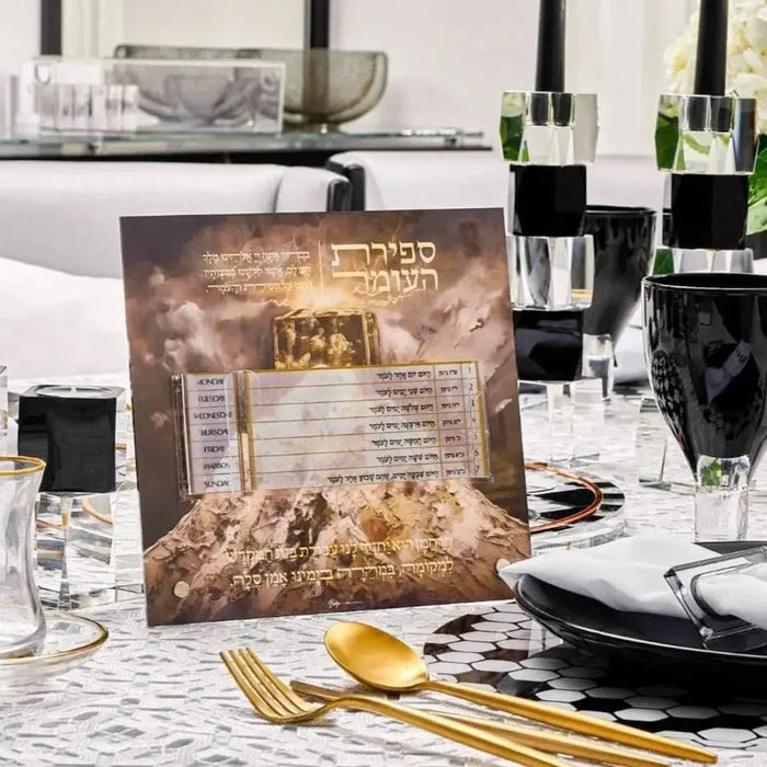 Tabletop Sefiras Haomer plaque by Batya with painted gold Jewish holiday design displayed on white patterned surface with gold cutlery and black dinnerware.