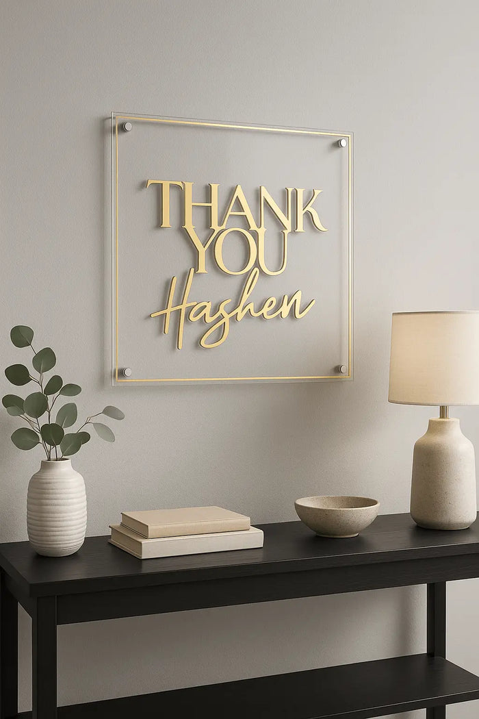 Thank You Hashem acrylic wall art in gold lettering on clear glass panel, displayed above black console table with decorative vases and books.