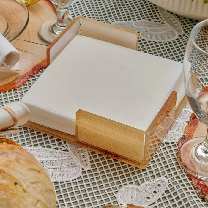 Natural wood napkin holder with white napkins displayed on a patterned tablecloth among place settings and tableware.