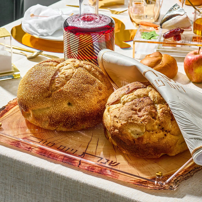 Wooden challah board with two round loaves of bread on a striped linen cloth, surrounded by Shabbat table settings and wine glasses.