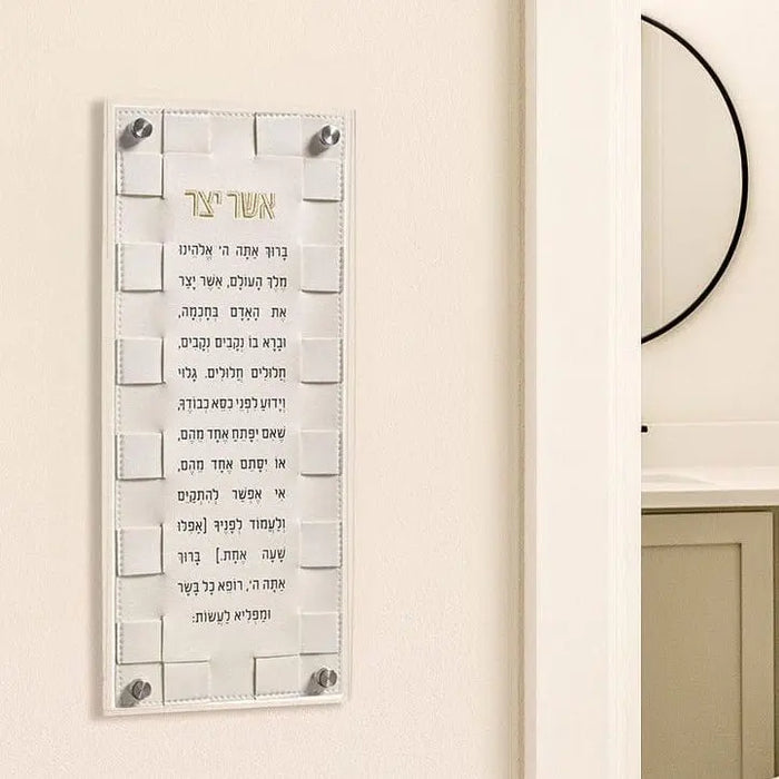 Woven Asher Yatzar wall art in cream with black text, displayed on beige wall with mounting hardware visible.