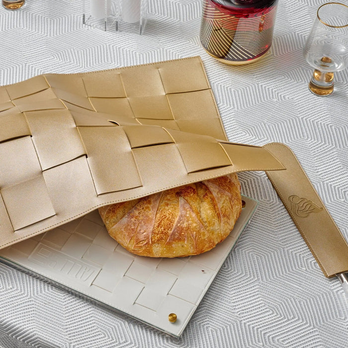 Gold woven challah board with a fresh baked challah loaf, displayed on white surface with Shabbat table setting elements.